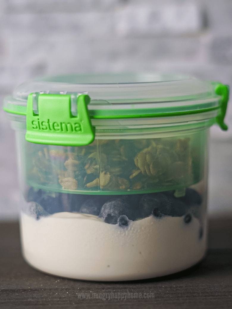 Closed travel parfait container with Greek yogurt and blueberries in the bottom and granola, chocolate ships and chia seeds in the top.