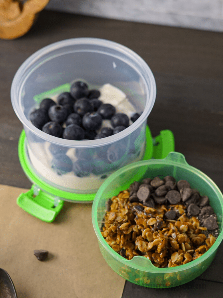 Open travel parfait container with Greek yogurt and blueberries in the bottom and granola, chocolate ships and chia seeds in the top.