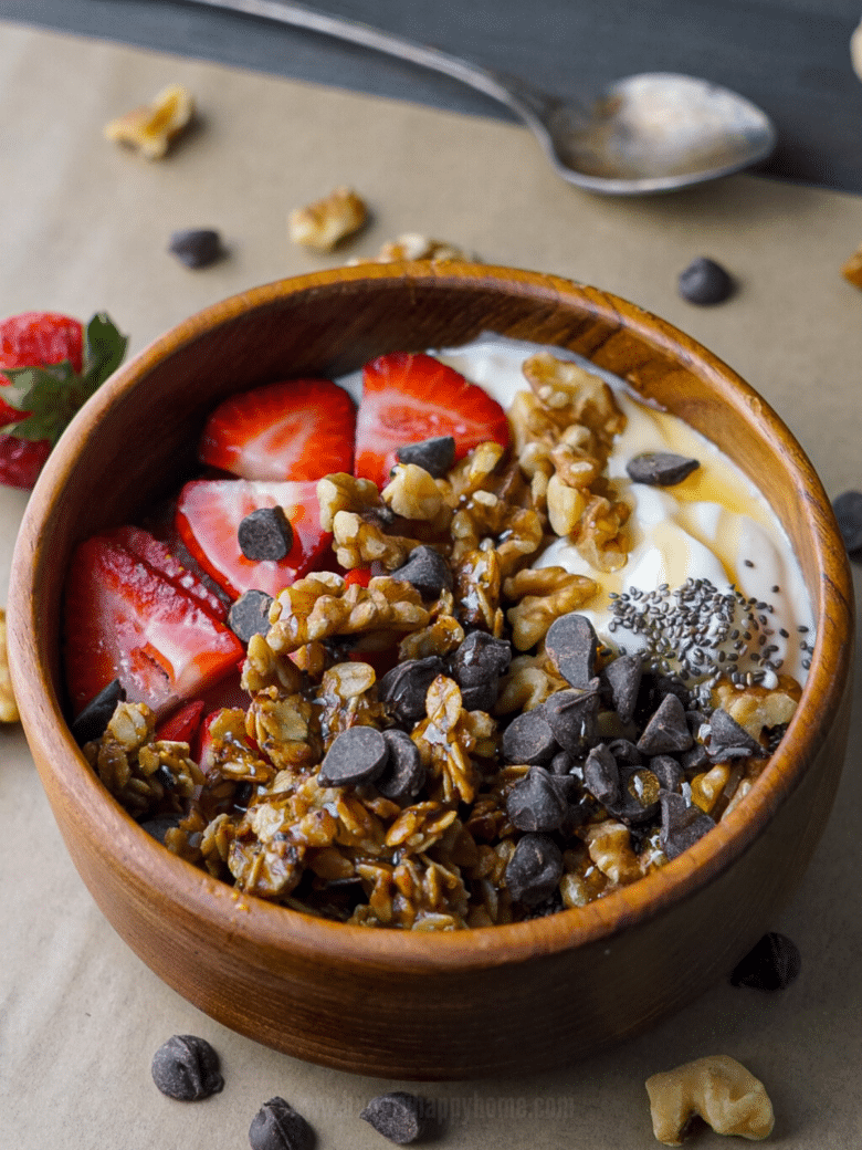 Wooden bowl with Greek yogurt topped with sliced strawberries, granola, walnuts, chocolate chips and chia seeds drizzled with honey.