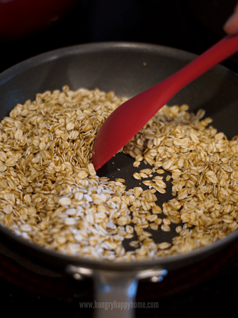 A red spatula stirring the toasting rolled oats in a pan on the stove.