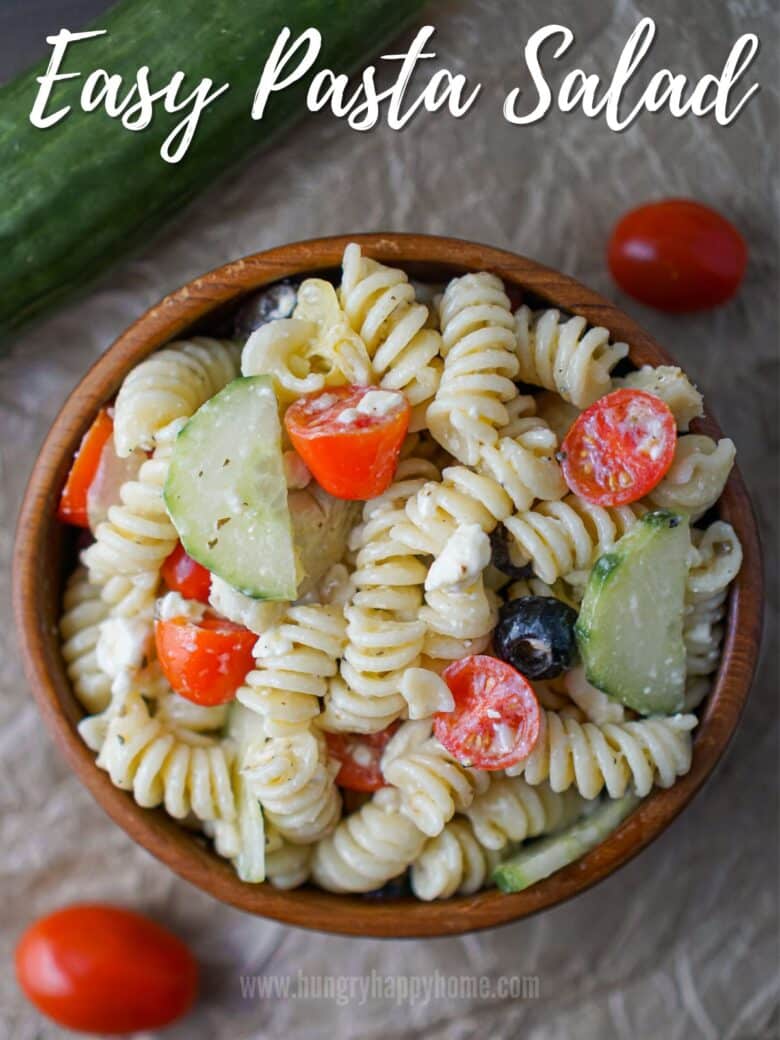 Wooden bowl with pasta salad in it that has cucumbers, tomatoes, olives and artichokes.