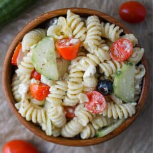 Wooden bowl with pasta salad in it that has cucumbers, tomatoes, olives and artichokes.